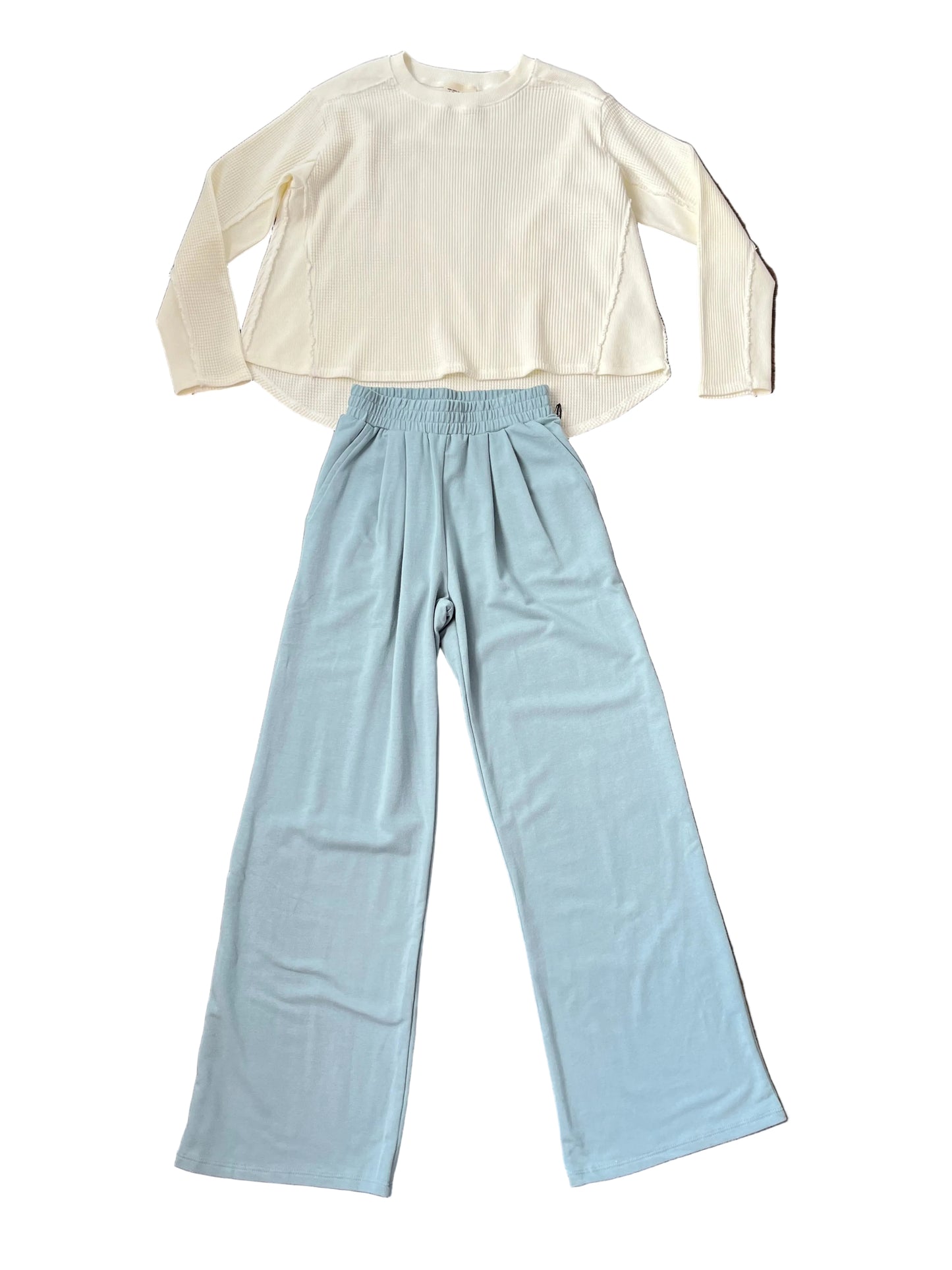 A two-piece clothing set laid flat on a dark wood floor, featuring a cream long-sleeve crop top and light blue wide-leg pants.