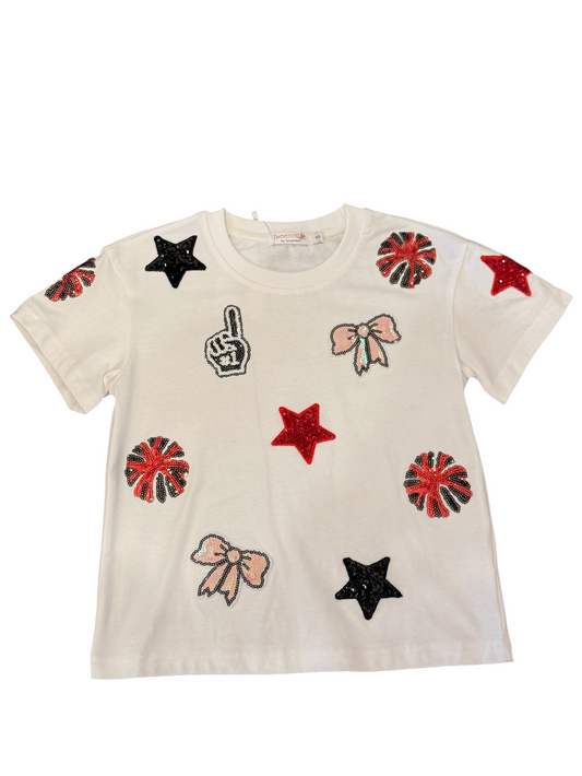 An off-white short-sleeve t-shirt for kids, decorated with various sequined patches including red and black stars, pink bows, and red-and-black pom-poms.