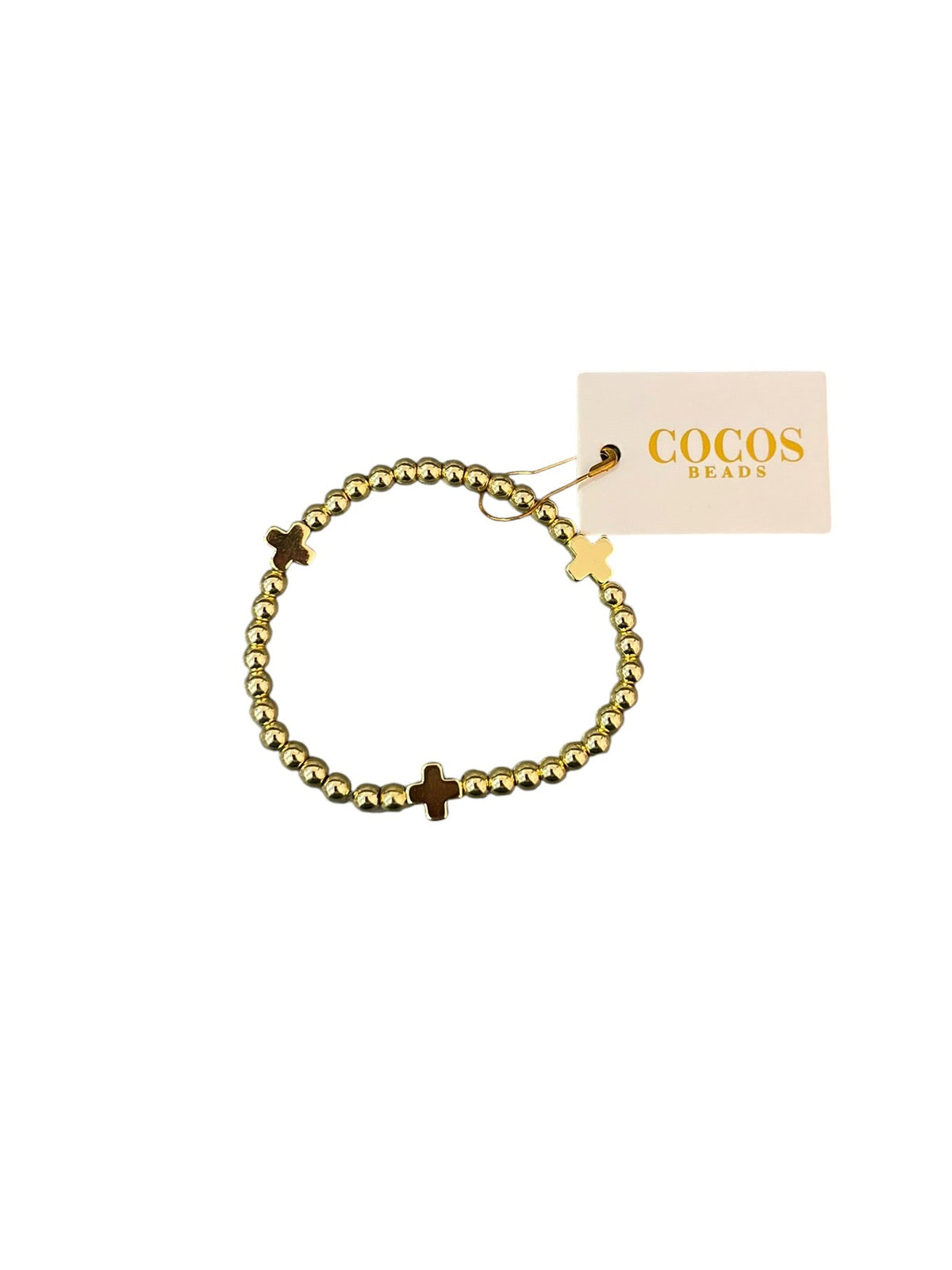 A gold beaded bracelet with three small gold cross charms, shown with its Cocos Beads brand tag on a white background.