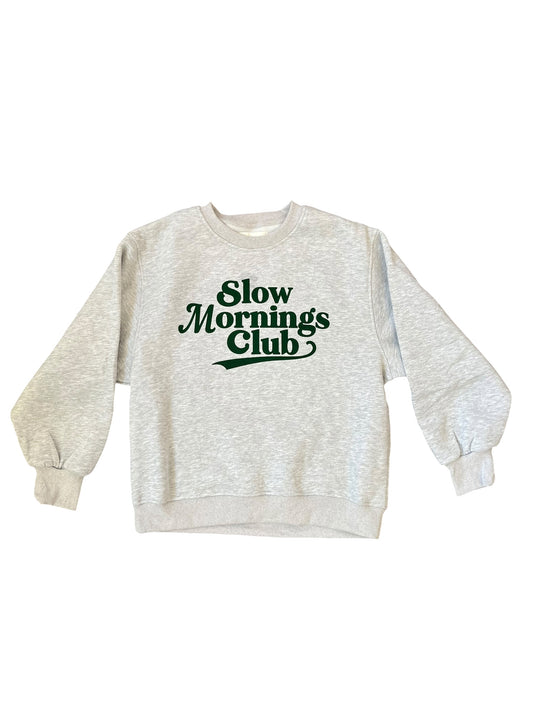 A light heather gray crewneck sweatshirt with 'Slow Mornings Club' printed in dark green across the chest, displayed flat against a white background.