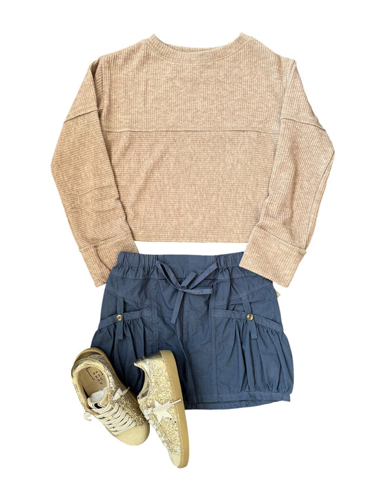A children's outfit flat lay featuring a beige ribbed knit long-sleeve crop top, navy blue ruffled skirt with a drawstring, and a pair of gold glitter sneakers, all arranged on a solid white background.
