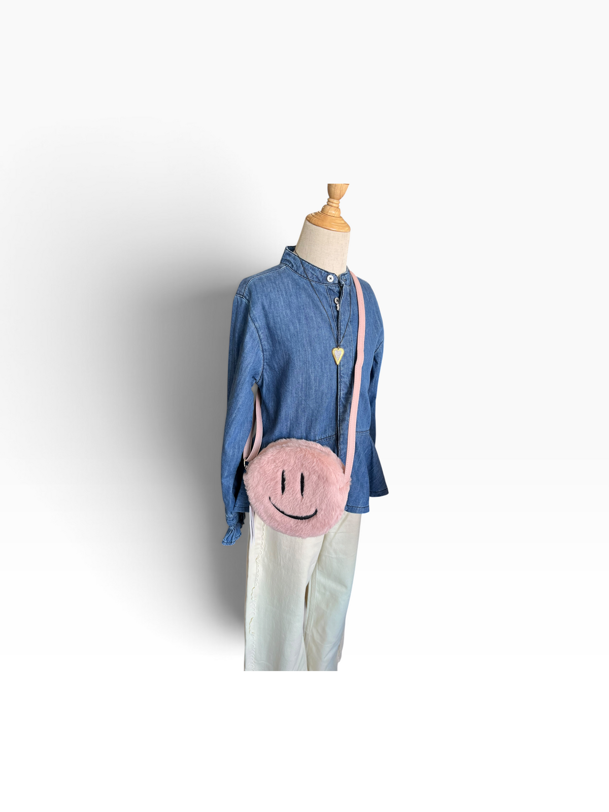 A pink fuzzy smiley face crossbody bag with a black embroidered face and a light pink strap, displayed on a mannequin wearing a blue denim shirt and white pants against a white background.